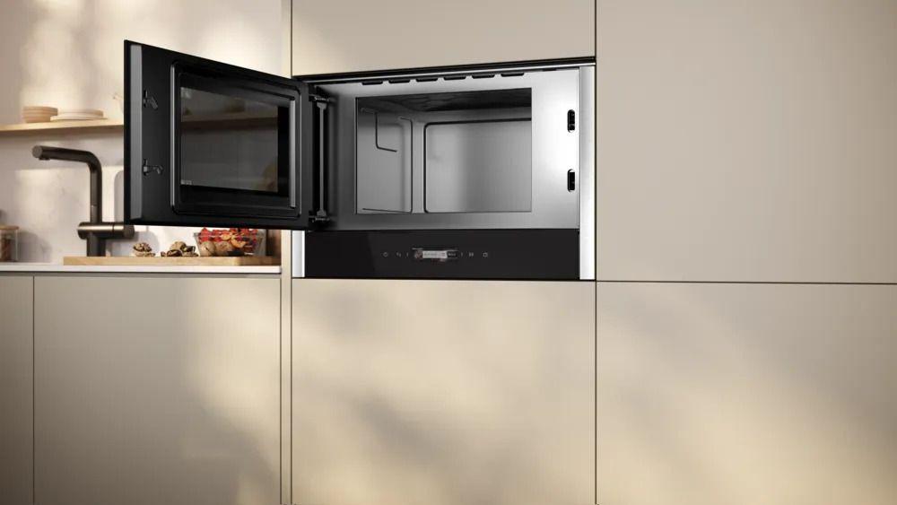 Eingebaute Mikrowelle mit geöffneter Tür in einer modernen, hellen Küchenfront mit flächenbündigem Design und schwarzem Wasserhahn im Hintergrund.